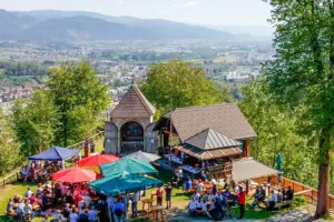 Viele Menschen auf Bänken vor einer Bergkapelle mit schöner Aussicht.