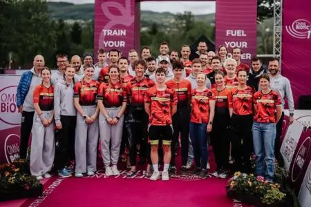 KSV-Triathlon kürte ihre Vereinsmeister am Stubenbergsee Eine große Gruppe von Triathletinnen, Triathleten und Betreuern posiert in Teamkleidung auf einer Eventbühne für ein gemeinsames Gruppenfoto. Im Hintergrund sind Sponsorenwände, Eventbanner und eine hügelige Landschaft zu sehen.