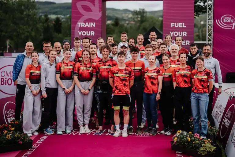 KSV-Triathlon kürte ihre Vereinsmeister am Stubenbergsee Eine große Gruppe von Triathletinnen, Triathleten und Betreuern posiert in Teamkleidung auf einer Eventbühne für ein gemeinsames Gruppenfoto. Im Hintergrund sind Sponsorenwände, Eventbanner und eine hügelige Landschaft zu sehen.