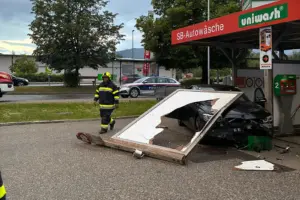 Ein beschädigtes schwarzes Auto steht an einer SB-Autowaschanlage, nachdem ein großes Werbeschild umgestürzt ist und auf das Fahrzeug gefallen ist. Ein Feuerwehrmann geht über den Parkplatz, im Hintergrund steht ein Polizeiauto an der Straße.