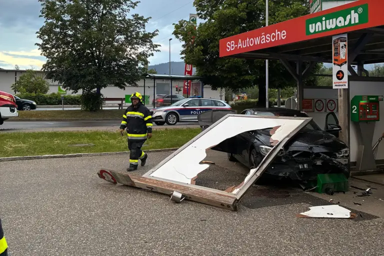 Kapfenberg: Auto krachte in Waschanlage Ein beschädigtes schwarzes Auto steht an einer SB-Autowaschanlage, nachdem ein großes Werbeschild umgestürzt ist und auf das Fahrzeug gefallen ist. Ein Feuerwehrmann geht über den Parkplatz, im Hintergrund steht ein Polizeiauto an der Straße.
