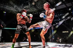 Zwei Kampfsportler stehen sich in einem beleuchteten MMA-Käfig gegenüber. Einer der Athleten setzt gerade einen Tritt an, während sein Gegner in Deckung geht und der Kampf von Zuschauern rund um die Arena verfolgt wird.