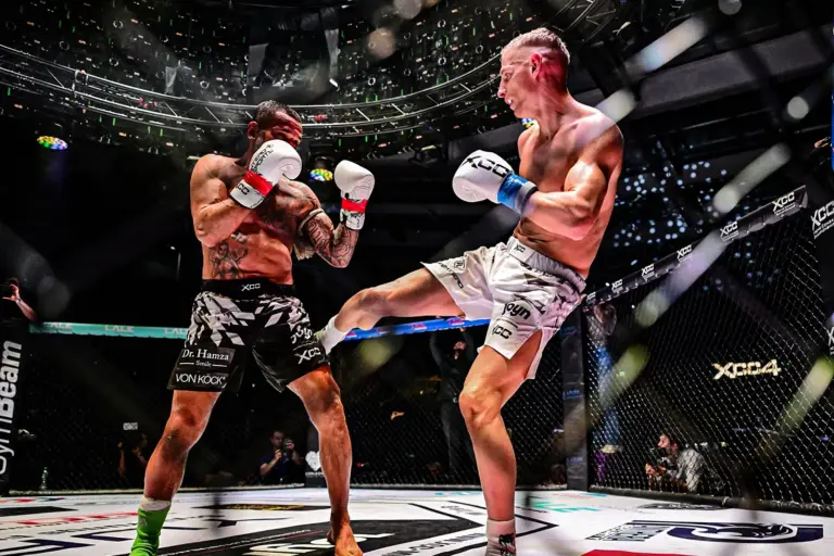 Idol zum Karriereabschluss k.o. geschlagen Zwei Kampfsportler stehen sich in einem beleuchteten MMA-Käfig gegenüber. Einer der Athleten setzt gerade einen Tritt an, während sein Gegner in Deckung geht und der Kampf von Zuschauern rund um die Arena verfolgt wird.