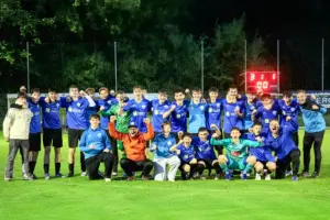 Eine Jugendfußballmannschaft in blauen Dressen jubelt nach einem Spiel gemeinsam auf dem Fußballplatz. Die Spieler und Betreuer posieren am Abend vor der Anzeigetafel mit erhobenen Fäusten für ein Gruppenfoto.