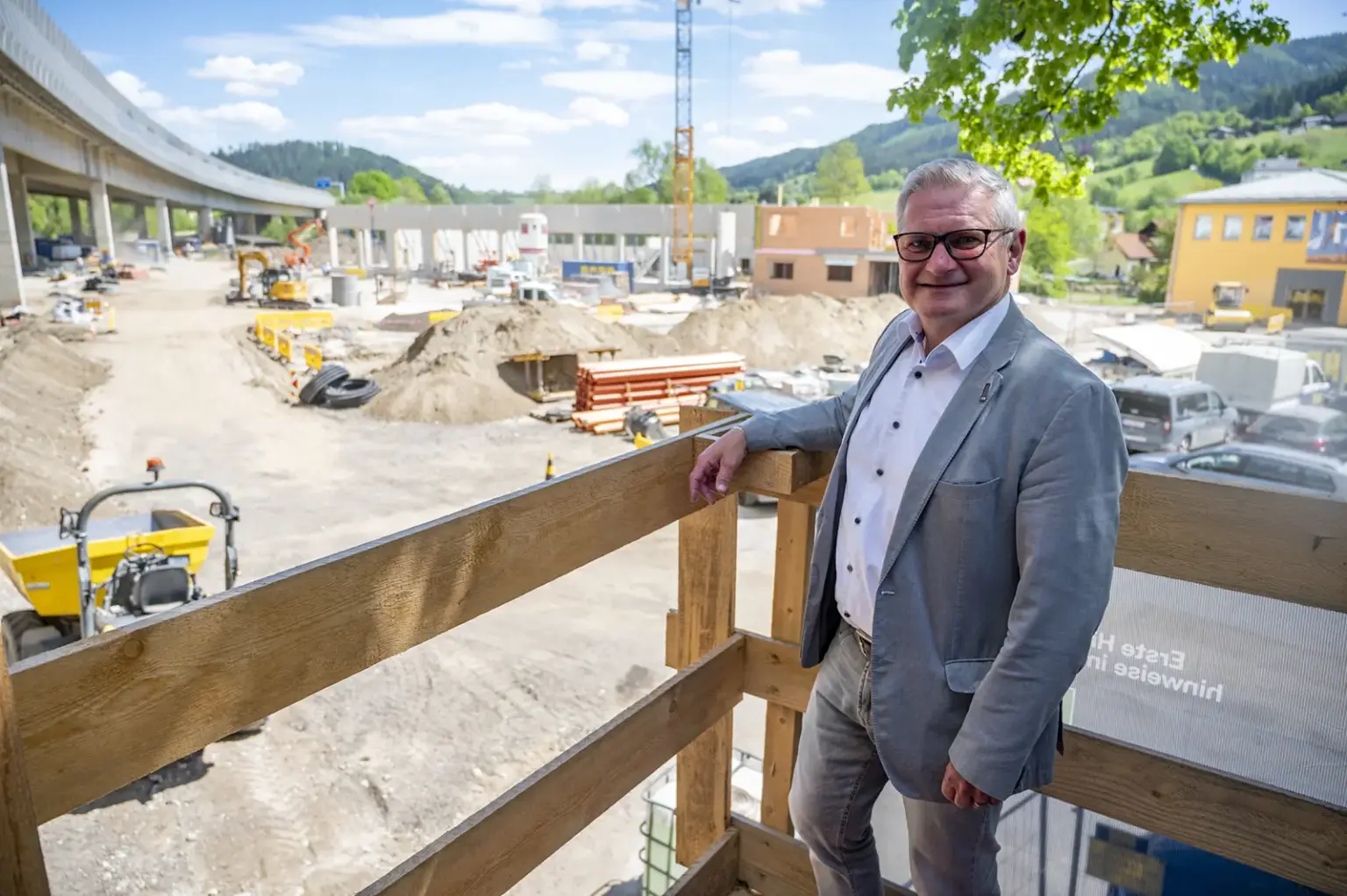Bau auf dem Roßdorfplatz schreitet zügig voran Ein Mann im Anzug steht auf einer Holzplattform vor einer großen Baustelle und blickt in die Kamera. Im Hintergrund sind Baugeräte, eine Brücke sowie Gebäude und Hügel in der Umgebung zu sehen.