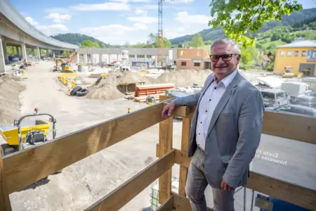 Ein Mann im Anzug steht auf einer Holzplattform vor einer großen Baustelle und blickt in die Kamera. Im Hintergrund sind Baugeräte, eine Brücke sowie Gebäude und Hügel in der Umgebung zu sehen.