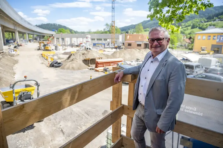 Bau auf dem Roßdorfplatz schreitet zügig voran Ein Mann im Anzug steht auf einer Holzplattform vor einer großen Baustelle und blickt in die Kamera. Im Hintergrund sind Baugeräte, eine Brücke sowie Gebäude und Hügel in der Umgebung zu sehen.