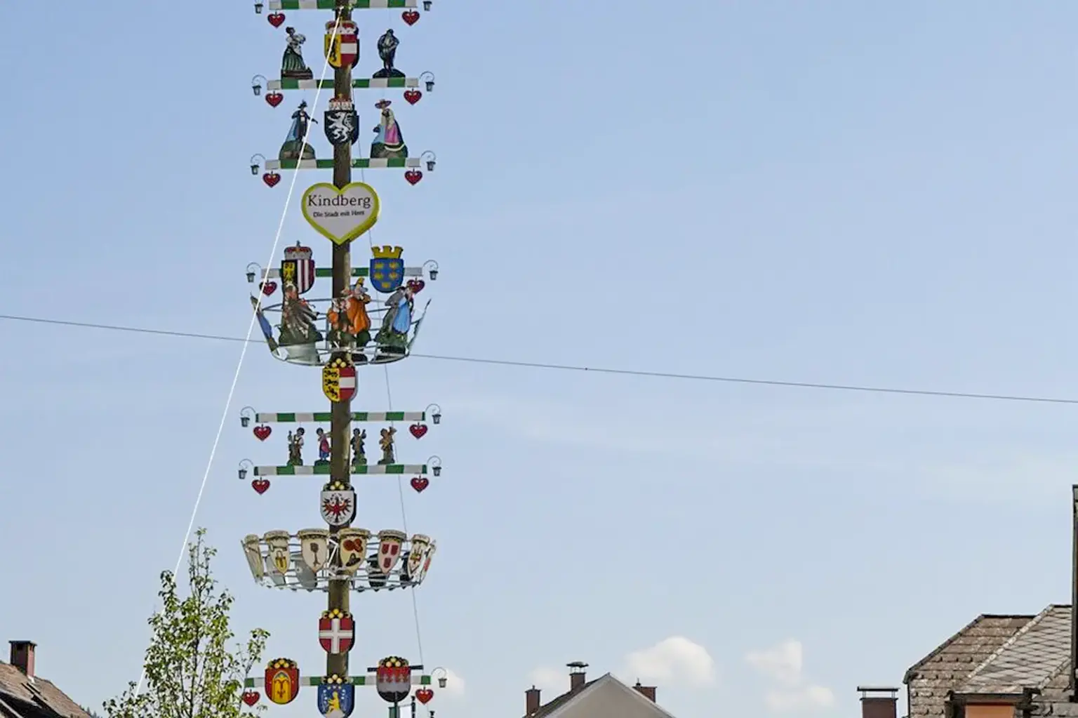 Zum Kindberger Kirtag steht der Zunftbaum wieder Ein festlich geschmückter Maibaum mit Wappen, Figuren und Herzen ragt vor einem hellen Himmel in die Höhe. Im unteren Bildbereich sind Dächer und Häuser einer Ortschaft zu sehen.