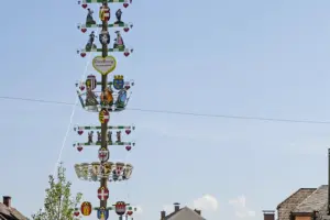 Ein festlich geschmückter Maibaum mit Wappen, Figuren und Herzen ragt vor einem hellen Himmel in die Höhe. Im unteren Bildbereich sind Dächer und Häuser einer Ortschaft zu sehen.