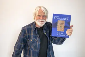 Ein älterer Mann mit weißem Haar und Bart hält ein blaues Buch mit dem Titel „Peter Rosegger & seine Gedanken“ in die Kamera. Er steht vor einer hellen Wand und lächelt freundlich.