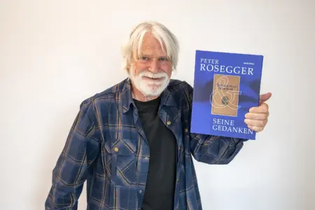 Ein älterer Mann mit weißem Haar und Bart hält ein blaues Buch mit dem Titel „Peter Rosegger & seine Gedanken“ in die Kamera. Er steht vor einer hellen Wand und lächelt freundlich.