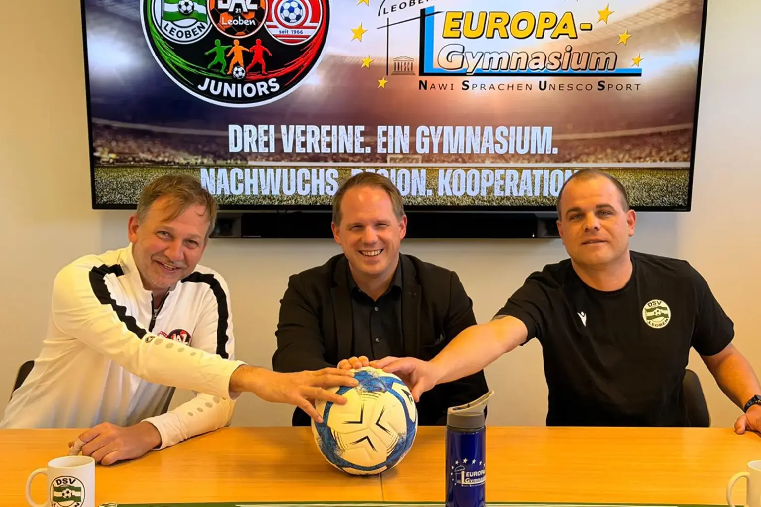 Drei Vertreter des Leobener Fußballs und des Europa-Gymnasiums sitzen an einem Tisch und legen gemeinsam ihre Hände auf einen Fußball – Symbol für eine neue Nachwuchskooperation. Im Hintergrund informiert ein Bildschirm über das gemeinsame Projekt von Vereinen und Schule.