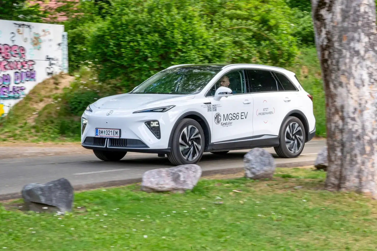 In der Ruhe liegt die Kraft Ein weißes Fahrzeug der Bauform SUV fährt in parkähnlicher Umgebung von links nach rechts, im Hintergrund eine mit Graffiti verzierte Mauer und Gebüsch
