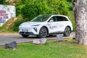 Ein weißes Fahrzeug der Bauform SUV fährt in parkähnlicher Umgebung von links nach rechts, im Hintergrund eine mit Graffiti verzierte Mauer und Gebüsch