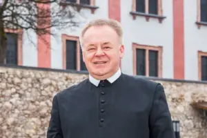 „In Mariazell entgeht man der Spiritualität nicht“ Porträt eines Geistlichen in schwarzer Soutane vor einem historischen Gebäude mit heller Fassade und Natursteinmauer. Der Mann blickt lächelnd in die Kamera.