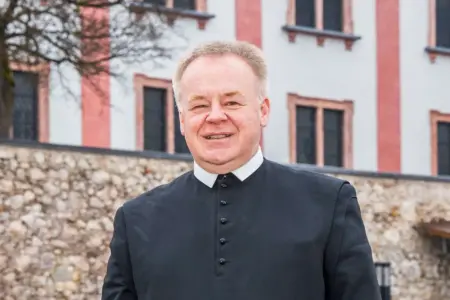 „In Mariazell entgeht man der Spiritualität nicht“ Porträt eines Geistlichen in schwarzer Soutane vor einem historischen Gebäude mit heller Fassade und Natursteinmauer. Der Mann blickt lächelnd in die Kamera.