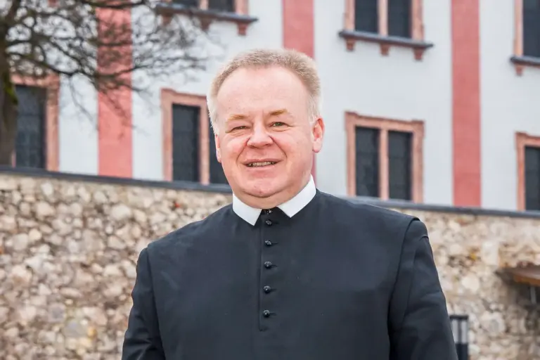 „In Mariazell entgeht man der Spiritualität nicht“ Porträt eines Geistlichen in schwarzer Soutane vor einem historischen Gebäude mit heller Fassade und Natursteinmauer. Der Mann blickt lächelnd in die Kamera.