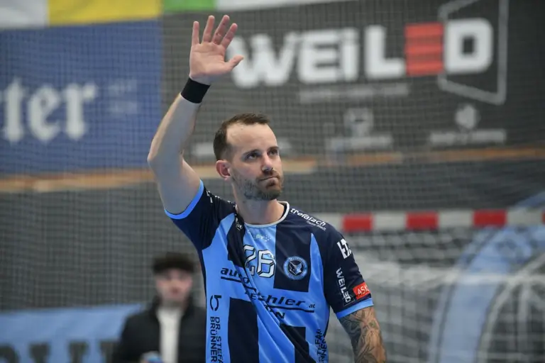 Ein Handballspieler in blau-schwarzem Trikot steht in einer Sporthalle vor dem Tor und hebt die rechte Hand zum Gruß. Er hat kurze Haare und einen Bart, trägt ein Armband am Handgelenk und blickt konzentriert nach oben. Im Hintergrund ist das Tor mit Netz sowie unscharfe Werbung zu sehen.