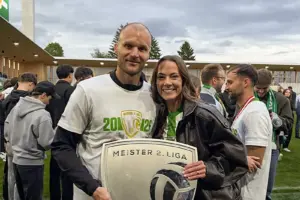 Ein Mann und eine Frau stehen nach einer Meisterfeier auf dem Fußballplatz nebeneinander und halten gemeinsam ein Meisterschild der 2. Liga. Im Hintergrund feiern weitere Personen im Stadion.