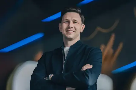 Mürzzuschlager wird Hoffenheim-Sportdirektor Ein Mann mit verschränkten Armen steht lächelnd vor einem dunklen Hintergrund mit blauen Lichtakzenten. Er trägt eine dunkle Jacke und blickt direkt in die Kamera.
