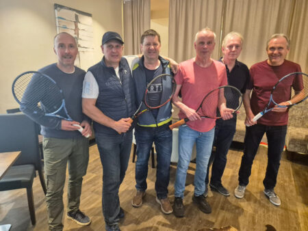 Sechs Männer stehen mit Tennisschlägern in einem Innenraum nebeneinander und posieren für ein Gruppenfoto. Die Gruppe lächelt in die Kamera, während einige der Männer ihre Schläger vor sich halten.