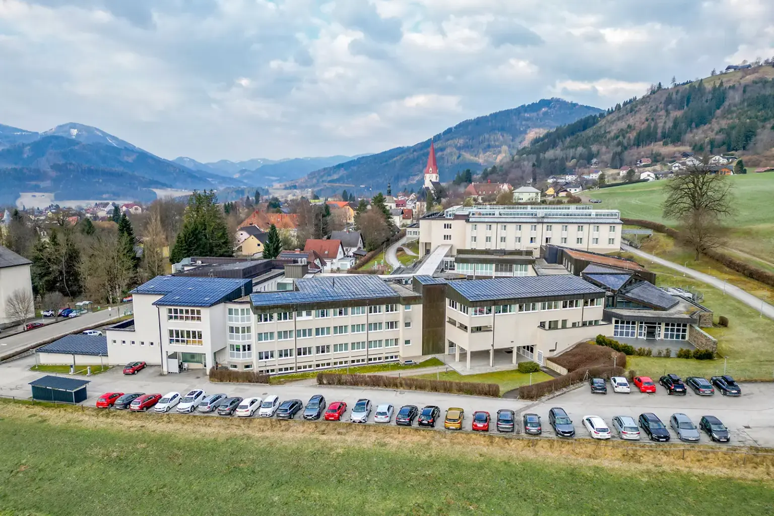 Landesparteien stehen geeint zum Rehazentrum Aflenz Ein größerer Gebäudekomplex in einer ländlichen Gegend, davor parkende Autos, im Hintergrund eine Ortschaft mit Kirchturm
