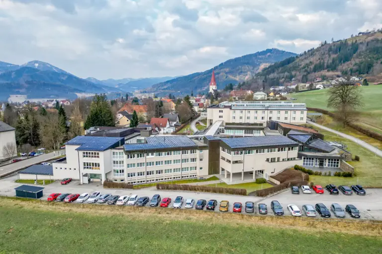 Ein größerer Gebäudekomplex in einer ländlichen Gegend, davor parkende Autos, im Hintergrund eine Ortschaft mit Kirchturm