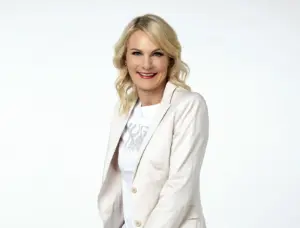 Eine blonde Frau im weißen Gewand schaut in die Kamera.