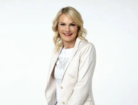 Eine blonde Frau im weißen Gewand schaut in die Kamera.