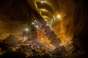 Ein schweres Bohr- und Abbruchgerät arbeitet tief im Inneren eines beleuchteten Eisenbahntunnels. Zwischen Felsbrocken und Staub treiben Maschinen den Tunnel Meter für Meter durch das Gestein.