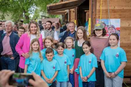 Gruppenfoto bei einer Sportlerehrung: Eine Frau im pinken Blazer posiert gemeinsam mit Kindern in hellblauen T-Shirts vor zahlreichen Besucherinnen und Besuchern im Freien.