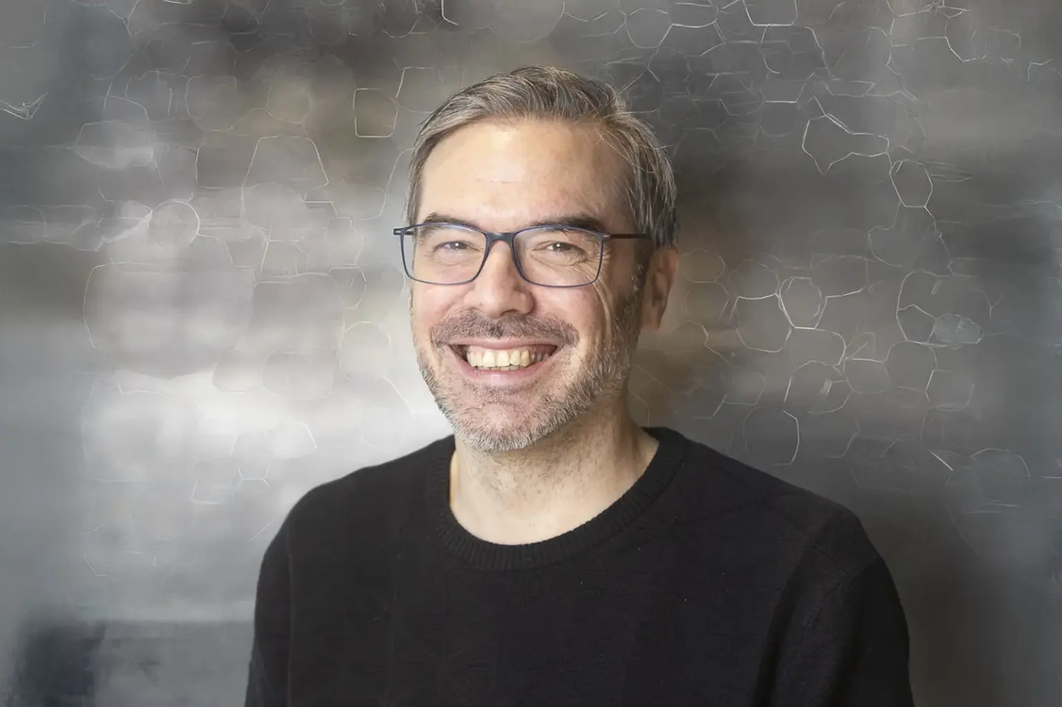 Porträt von Thomas Baumann: Der Mann mit Brille und kurzem grauem Haar lächelt in die Kamera; aufgenommen vor einem dezent strukturierten, unscharfen Hintergrund.