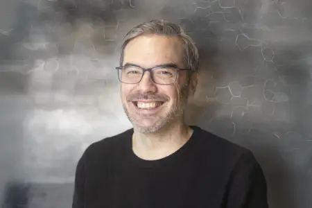 Porträt von Thomas Baumann: Der Mann mit Brille und kurzem grauem Haar lächelt in die Kamera; aufgenommen vor einem dezent strukturierten, unscharfen Hintergrund.