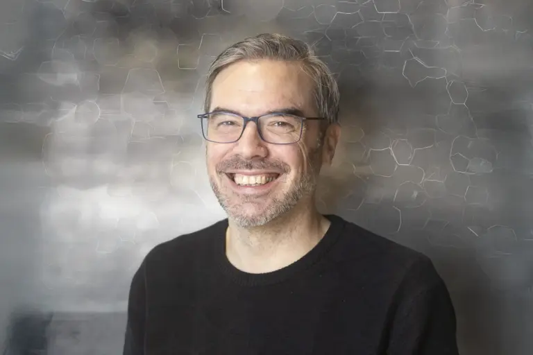Porträt von Thomas Baumann: Der Mann mit Brille und kurzem grauem Haar lächelt in die Kamera; aufgenommen vor einem dezent strukturierten, unscharfen Hintergrund.