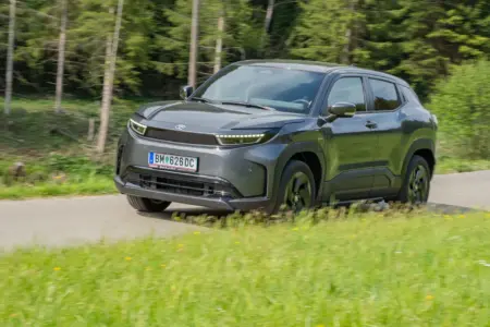 Ein grauer PKW der Kategorie Kompakt-SUV fährt auf einer Landstraße von rechts nach links, im Vordergrund eine Wiese, im Hintergrund Wald