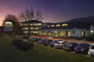 Das WIFI Niklasdorf ist in der Abenddämmerung beleuchtet zu sehen. Vor dem Gebäude stehen mehrere geparkte Autos, im Hintergrund sind Bäume und die Silhouette der umliegenden Berge erkennbar.