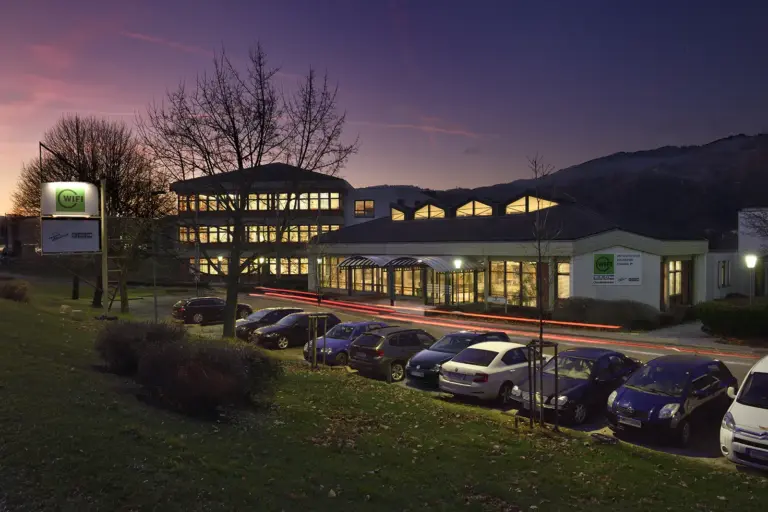 Niklasdorf wird zum WIFI-Holzstandort Das WIFI Niklasdorf ist in der Abenddämmerung beleuchtet zu sehen. Vor dem Gebäude stehen mehrere geparkte Autos, im Hintergrund sind Bäume und die Silhouette der umliegenden Berge erkennbar.