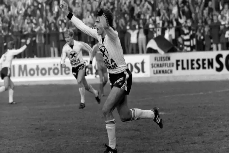 chwarz-Weiß-Aufnahme eines Fußballspiels: Ein Spieler sprintet mit jubelnd erhobenen Armen über den Platz, während im Hintergrund Mitspieler und Fans den Treffer feiern.