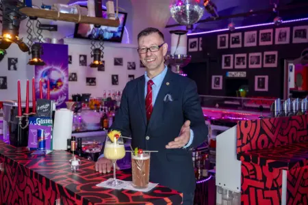 Afoch guat: Italienische und asiatische Grüße im Cocktailglas Ein Mann im Anzug steht hinter einer Bar und hält eine Hand einladend nach vorne. Vor ihm stehen zwei dekorierte Cocktails auf der Theke, im Hintergrund sind Barregale, eine Discokugel und farbige Beleuchtung zu sehen.