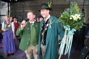 Zwei Männer in Trachtenkleidung stehen auf einer Bühne, der linke spricht in ein Mikrofon, der rechte hält ein Gebinde aus Grass und Blumen.