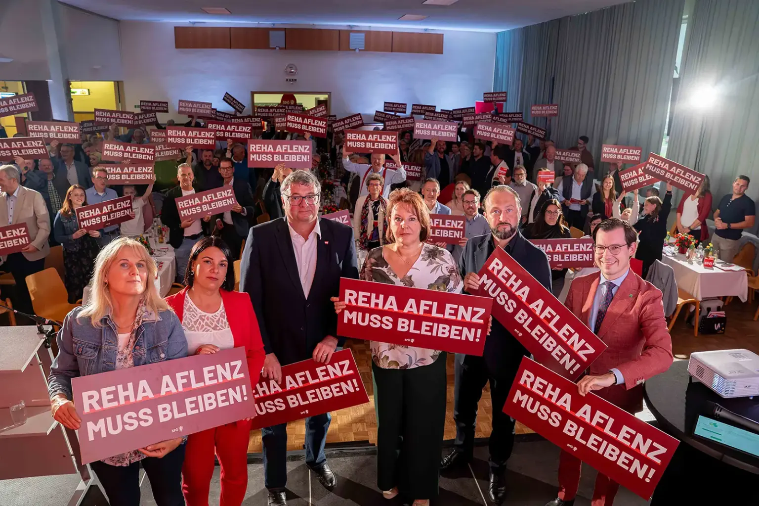 Rote Bezirkskonferenz in Thörl Menschen in einem Saal mit Schildern in Händen, darauf die Aufschrift "Reha Aflenz muss bleiben!"