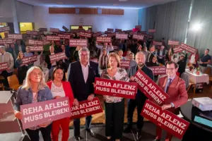 Menschen in einem Saal mit Schildern in Händen, darauf die Aufschrift "Reha Aflenz muss bleiben!"