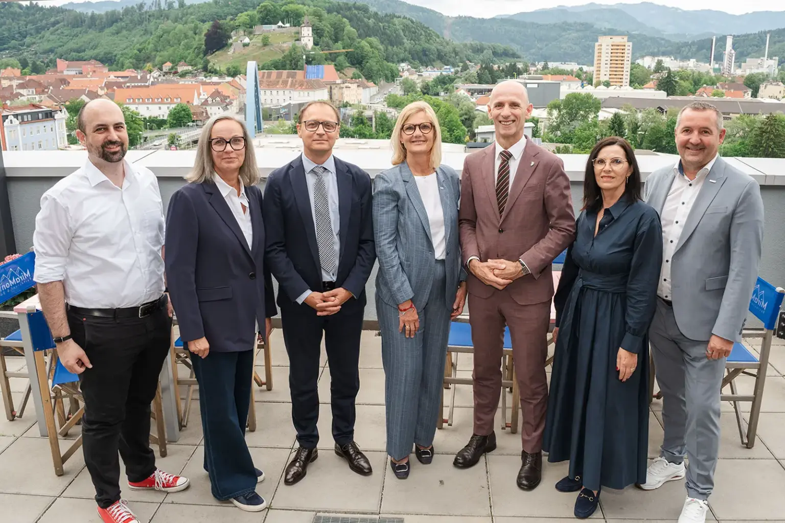 Drei Frauen und vier Männer stehen auf einer Terrasse, im Hintergrund eine Stadt