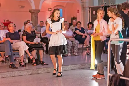 Drei junge Frauen in Kostümen stehen vor einem älteren Publikum