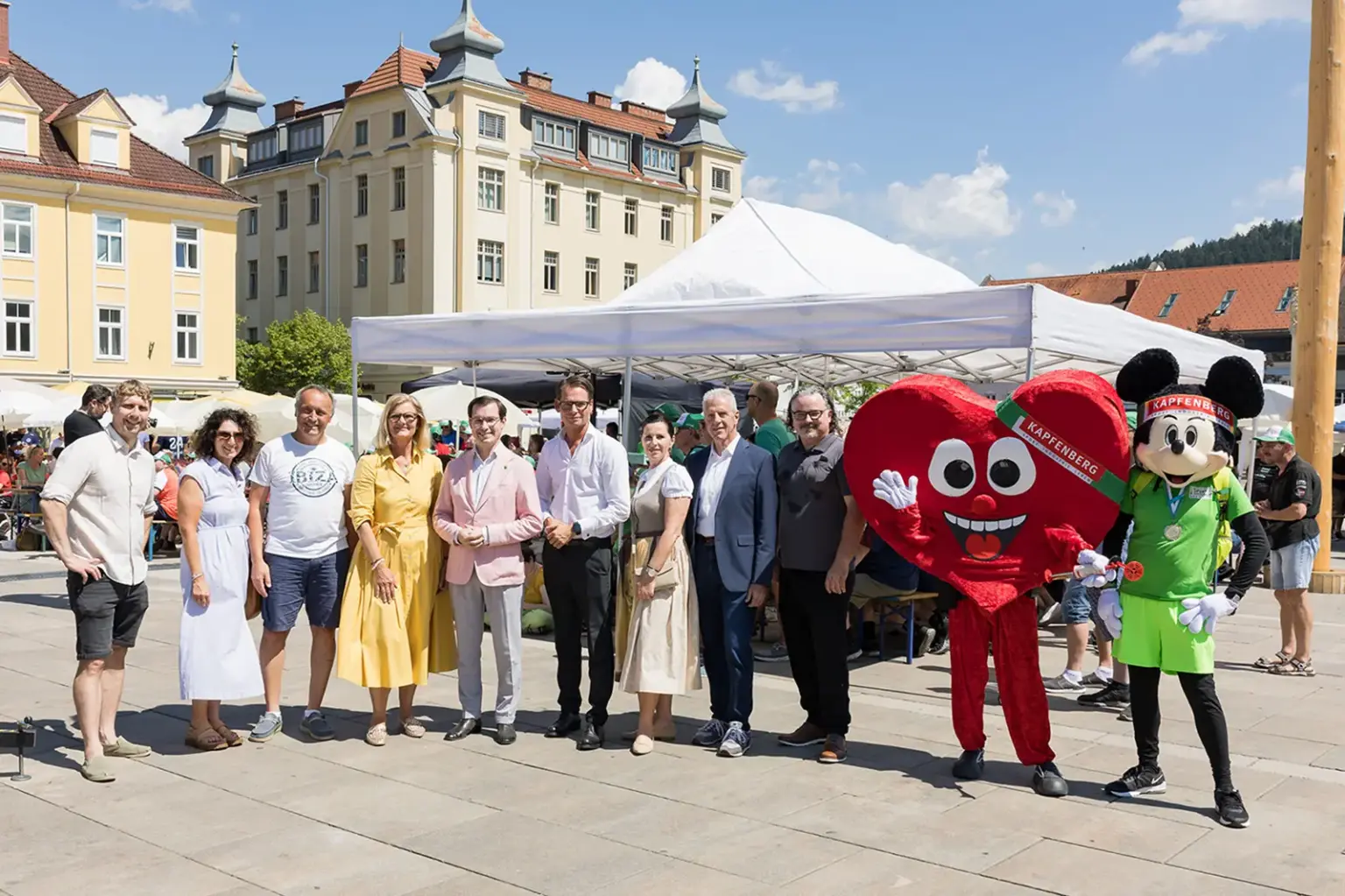 Bruck: Abschied für die steirischen Athleten Eine Gruppe von Personen steht mit zwei Maskottchen auf einem großen Platz vor einem Pavillon