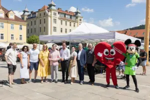 Eine Gruppe von Personen steht mit zwei Maskottchen auf einem großen Platz vor einem Pavillon