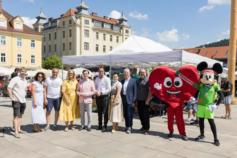 Bruck: Abschied für die steirischen Athleten Eine Gruppe von Personen steht mit zwei Maskottchen auf einem großen Platz vor einem Pavillon