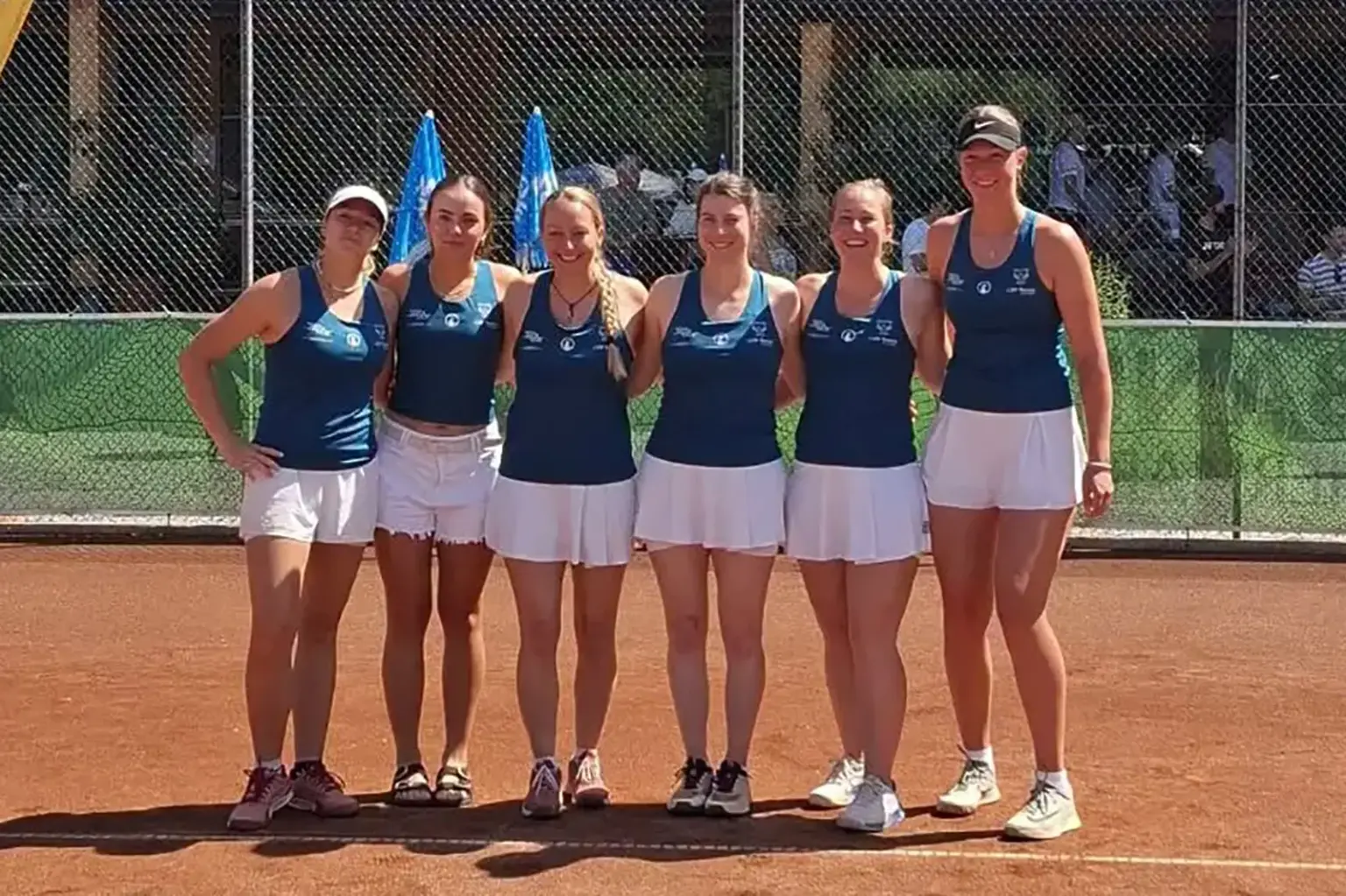 Damenmannschaft posiert vor einem Meisterschaftsspiel auf einem Tennis-Sandplatz für ein Gruppenfoto.