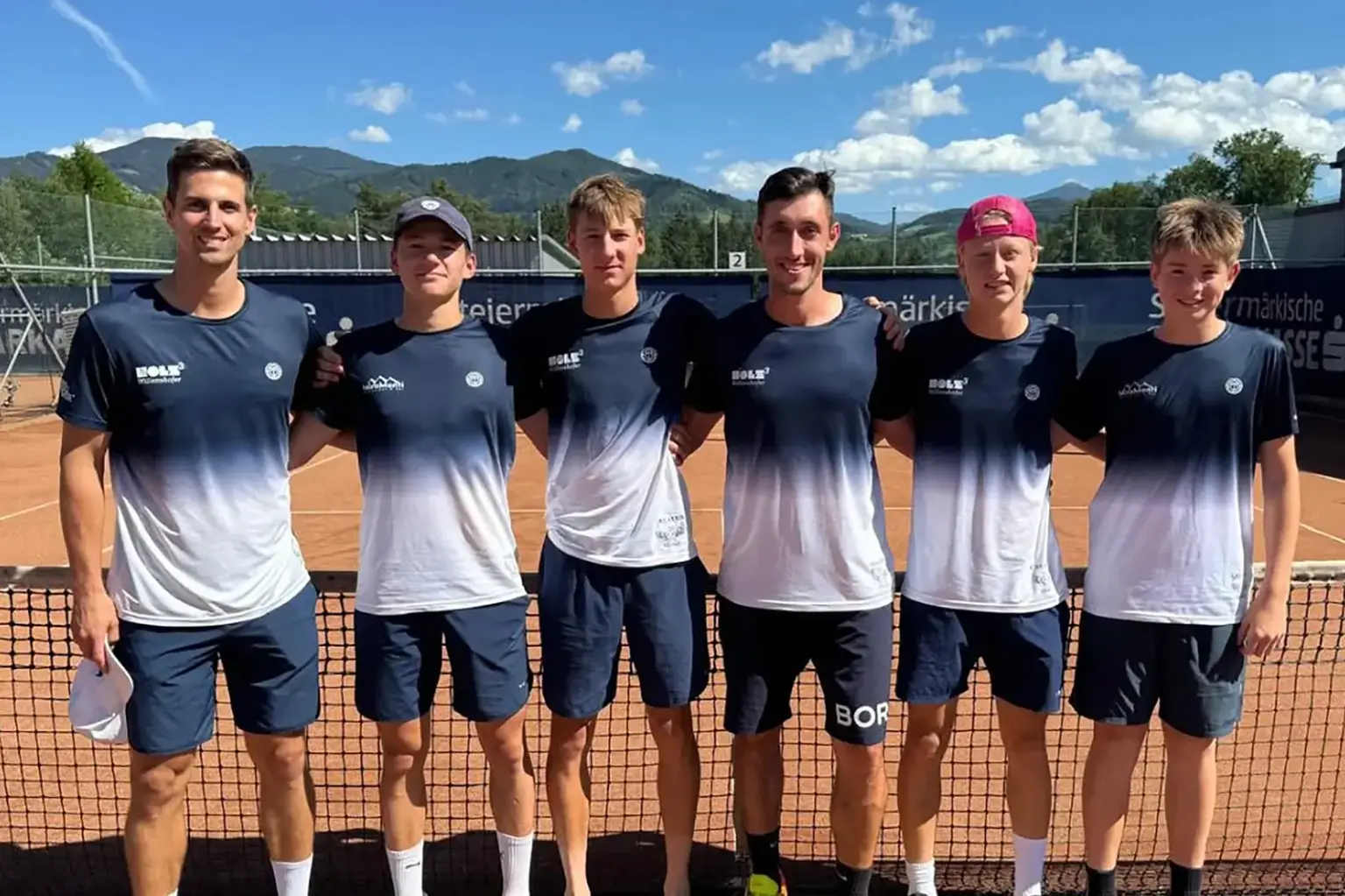 Sechs Tennisspieler stehen auf einem Sandplatz vor dem Netz und posieren für ein Mannschaftsfoto. Im Hintergrund sind Berge, blauer Himmel und die Tennisanlage zu sehen.