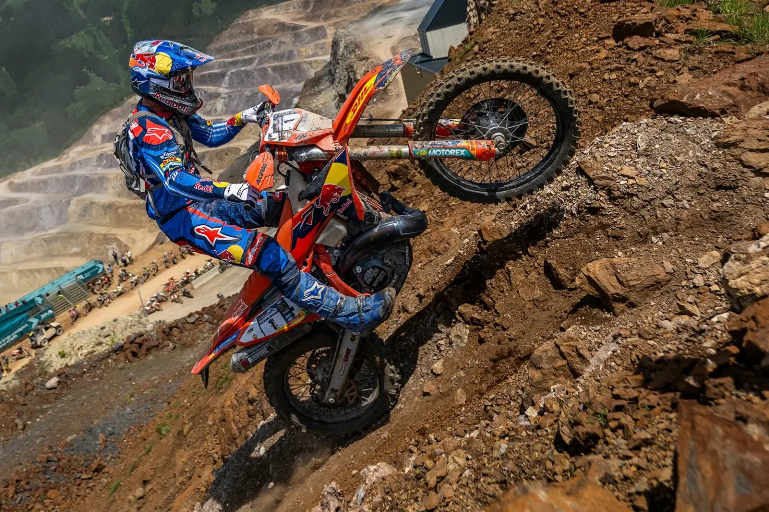 Erzbergrodeo: Fünfter Streich zum Jubiläum Ein Enduro-Motorradfahrer in blau-roter Schutzausrüstung kämpft sich eine extrem steile, felsige Abraumhalde hinauf. Das Vorderrad des orangefarbenen Motorrads hebt sich über eine Geländekante, während Geröll unter den Reifen wegrutscht. Im Hintergrund sind die terrassenförmigen Stufen eines großen Tagebaus sowie weitere Motorräder und Zuschauer zu erkennen. Die Aufnahme vermittelt Dynamik, Kraft und die Herausforderung des anspruchsvollen Offroad-Wettbewerbs.
